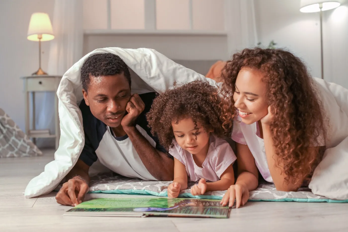 Parents reading a picture book with their young daughter under a blanket fort